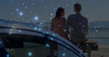 Okyanus kenarında bankta oturan çift, parlayan mavi animasyon parçacıklarını izliyor. Romantik, akşam, sükunet, açık hava