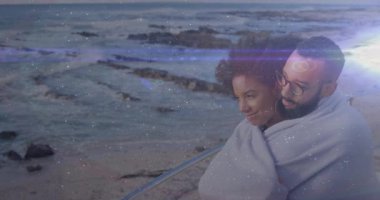 Battaniyeye sarılmış, plajda ışık efektleriyle animasyon izleyen bir çift. Romantizm, sarılma, akşam, sıcak, açık hava, boş zaman