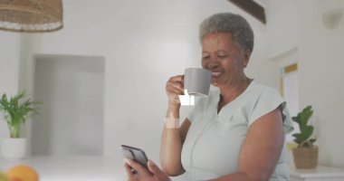 Akıllı telefondan hoşlanan, evde kahve içen yaşlı bir kadın. Son sınıf, teknoloji, iletişim, rahatlama, yaşam tarzı.