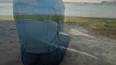 Kot ceket giymiş, okyanus dalgalarına ve manzaralı tarlalara bakan biri. Doğa, manzara, sükunet, tefekkür, özgürlük, macera