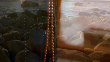 Elinde boncuklar, arka planda okyanus dalgalarıyla kayalık sahilde duran bir kadın. Meditasyon, sükunet, doğa, dinginlik, huzur