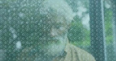 Yağmurla kaplı camların ardında sakallı yaşlı bir adam sessizce dışarı bakıyor. Düşünce, yalnızlık, sakal, nostalji, huzur, yansıma.