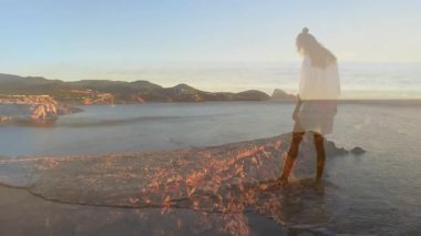 Kayalık kayalıklarda duran, sakin okyanusa ve uzak tepelere bakan kadın animasyonu. Macera, sükunet, keşif, seyahat, manzara, doğa