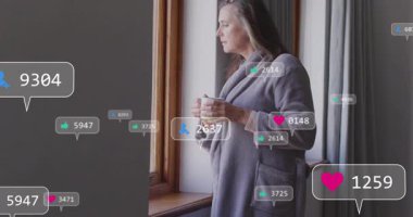 Elinde kupa tutan kadın, pazarlama ölçülerini gösteren yüzen sosyal medya simgeleriyle pencereden bakıyor. Düşüncelere dalma, rahatlama, ev, modern, dijital, konfor, yaşam tarzı