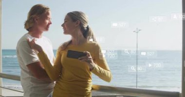 Kadın akıllı telefonu tutarken erkekler okyanus korkuluklarına yaslanıyor pazarlama ölçüm simgeleri sergiliyor. Romantizm, sosyal paylaşım, açık hava, manzara, teknoloji, bağlantı, mutluluk