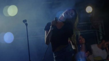 Smoky müzik sahnesinde mikrofon çeken solist, mavi spot ışıkları altında dans eden animasyon ses dalgaları. Konser, performans, müzik, gece hayatı, sahne, aydınlatma, grup