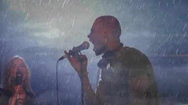 Erkek şarkıcı, yağmura boğulmuş sahnede müzik notalarıyla seyirciye doğru uzanıyor. Performans, açık hava, konser, dinamik, duygusal, canlı, işbirliği