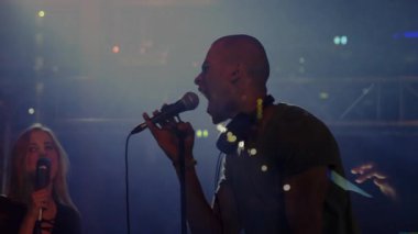Sahne ışıkları altında, animasyon müzik ikonlarıyla şarkı söyleyen mikrofon ve akıllı telefon tutan iki şarkıcı. Performans, eğlence, canlı müzik, vokalistler, konser, sahne, gece hayatı