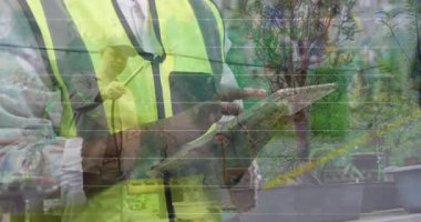 Bahçıvan bitkileri finansal haritalarla sularken, Hi-vis işçi tableti tıkırdatıyor. Endüstri, ilerleme, yenilik, teknoloji, bahçıvanlık, manzara, büyüme