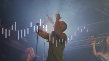 Loş ışıkta sahne alan üç şarkıcı, finans grafikleri ve pembe trend örtüsünü harmanlıyor. Performans, konser, vokalistler, sahne, müzik, gece kulübü, eğlence
