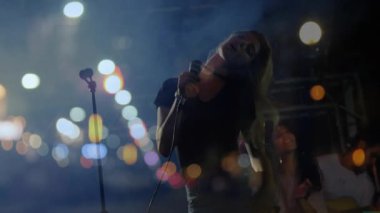 Mikrofon tutan kadın şarkıcı, ışıklar altında müzik yapan şehir bataklığı arkasında yükselen ses parmaklıkları. Canlı müzik, performans, gece, şehir, konser, sahne, eğlence