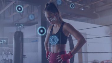 Kadın boksör kum torbasının yanında duruyor, kırmızı el ambalajları takıyor, holografik fitness HUD gösteriyor. Atletik, spor salonu, fütüristik, eğitim, güçlendirme, güç, dijital