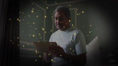 Son sınıf öğrencisi yatak odasında tablet kullanıyor. Parlayan parçacıklar etrafta uçuşuyor ve teknoloji temasını gösteriyor. Yaşlılık, rahatlama, sağIık, huzur, ortam, dijital, konfor