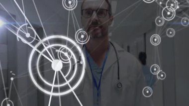 Hastane koridorunda duran doktor dairesel HUD düğümlerini gösteriyor ve tıbbi veri hatlarını birbirine bağlıyor. Tıbbi, sağlık, teknoloji, yenilik, analiz, fütüristik, profesyonellik