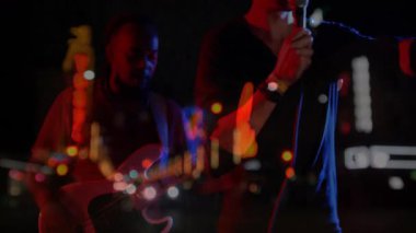 Şarkıcı mikrofon tutuyor ve gitarist, neon müzik efektleriyle sokak gecesinde gitar çalıyor. Konser, performans, gece hayatı, şehir, canlı, sanatsal, eğlence