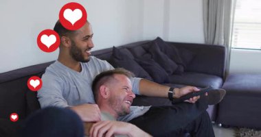 İki adam televizyon kumandası ve yüzen kalp ikonlarıyla ev eğlencesi sahnesinde kanepede sarılıyor. Romantizm, samimiyet, arkadaşlık, modern, samimi, sevecen, dostluk