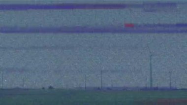 Çit direkleri ve türbin silueti ile noktalanmış düz çimenli alan görünüyor. Manzara, distopyan, sessiz, atmosferik, endüstriyel, kırsal, dijital gürültü