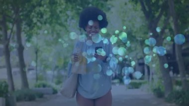 Şehir yolunda yürüyen bir kadın çantasıyla akıllı telefona bakıyor, yüzen teknoloji grafikleri gösteriyor. Kentsel, yaşam tarzı, gündelik, modern, açık hava, sağlık, teknoloji