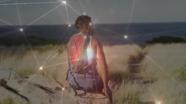 Kayaların üzerinde oturan adam okyanus yoluna bakan hidrasyon paketi takıyor, teknoloji ağı üst üste biniyor. Doğalcı, maceracı, keşif, fitness, açık hava, enerjik, minimalist