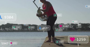 Tahta iskelede diz çökmüş bir kadın göl kenarında yüzen fitness medya simgelerini gösteriyor. Sporcular, rahatlama, susuzluk, doğa, açık hava aktivitesi, zindelik, zindelik