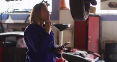 Tamirhanede kaldırılmış araçları inceleyen kadın tamirci, animasyon tablet verileri ve lens fişeğiyle. Profesyonel, endüstriyel, enerjik, yenilik, bakım, teknoloji, takım çalışması