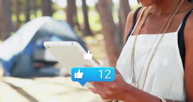 Orman kamp alanında tablet tutan bir kadın, yüzen sosyal medya bilgilendirme simgeleri sergiliyor. Doğa, açık hava, teknoloji, eğlence, macera, yaşam tarzı, sükunet