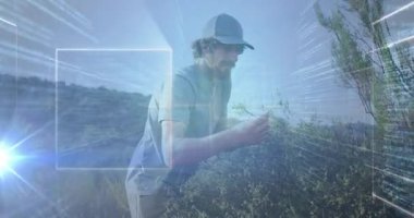 Kuru çalıların arasında çömelmiş bir adam, yüzen teknoloji kaplamalarıyla bitki sağlığını ve mercek parlamasını inceliyor. Açık hava, teknoloji, macera, keşif, yenilik, dayanıklı, fütürist