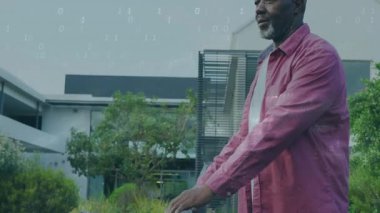 Olgun bir adam ön bahçede öne eğiliyor, yüzen dijital ikili sistemle teknolojiyi gösteriyor. Düşünceli, modern, minimalist, profesyonel, sakin, kentsel, yaratıcı