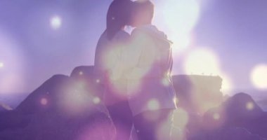 Kayalık sahilde sarılıp öpüşen çift pastel bokeh fişekleri ile pazarlama grafiklerini gösteriyor. Romantizm, bağlantı, samimiyet, açık hava, manzara, hassas, yumuşak