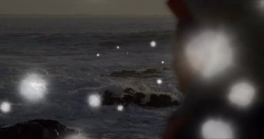 Sağlık animasyonunda farkındalığı keşfeden bir kadın, dalgalar ve yüzen bokeh ışıkları. Deniz manzarası, sükunet, meditasyon, alacakaranlık, engebeli, ruhani, içgözlem