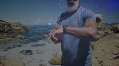 Olgun bir adam Rocky Sahili 'nde animasyon fitness grafikleri ve yüzen veri simgeleriyle akıllı saatleri kontrol ediyor. Açık hava, atletik, minimalist, sağlık, peyzaj, konsantrasyon, teknoloji