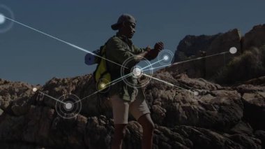 Kayalık çıkıntıda duran erkek yürüyüşçü dijital ağ düğümlerini gösteren akıllı kameraları kontrol ediyor. Macera, keşif, açık hava, hareketlilik, teknoloji, bağlantı, dayanıklılık