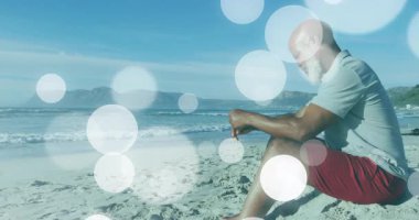 Yaşlı adam kumlu sahilde oturuyor, sağlık meditasyonunu gösteren animasyon ışıkları var. Rahatlama, tefekkür, dinginlik, rahatlık, kıyı, ufuk, gün batımı.