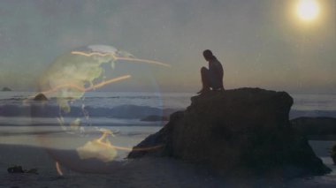 Gün batımında plaj kayasında oturan kadın silueti teknoloji ağı ile kaplanmış küreyi gösteriyor. Siluet, sükunet, yenilik, bağlantı, keşif, doğa, sükunet