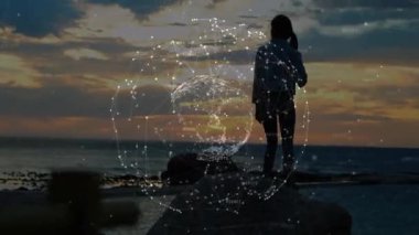 Gün batımında kıyı kayasında duran bir kadın teknoloji ağı kürelerini sergiliyor. Macera, keşif, bağlantı, esneklik, modern, açık hava, manzara