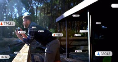 Ahşap korkuluklara yaslanmış bir adam yüzen sosyal medya ikonlarıyla ormanı yakalıyor. Doğa, macera, teknoloji, açık hava, keşif, gerçeklik, sükunet