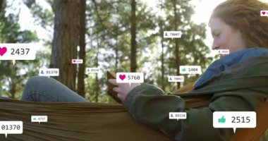 Ormanda hamakta uzanan bir kadın akıllı telefonuyla etkileşime geçiyor, sosyal medya simgeleri etrafta dolanıyor. Doğa, rahatlama, dijital, bağlantı, açık hava, rahatlık, huzur