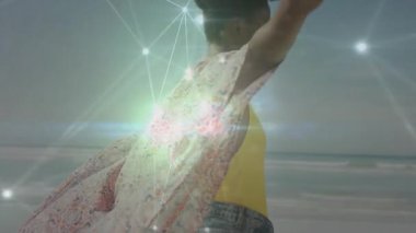 Kadın sahilde dengede duruyor, renkli şallar sergiliyor, yüzen şebeke çizgileri teknik bağlantıyı gösteriyor. Bohem, canlı, dijital, dinginlik, açık hava, anlamlı, enerjik