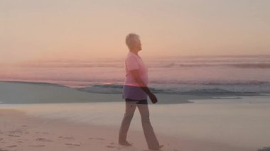 Kıyı şeridi boyunca yürüyen yaşlı kadın, canlandırılmış kalp ikonu gösteriyor ve sağlık uygulamaları için adımları sayıyor. Deniz kenarı, sağIık, huzur, ufuk, pastel, yansıma, rahatlık