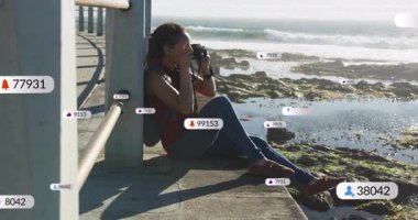 Kadın elinde kamerayla okyanus kıyısındaki iskelede yüzen sosyal medya pazarlama ikonlarıyla okyanusu çekiyor. Çağdaş, kıyı, açık hava, macera, yaşam tarzı, belgesel, canlı