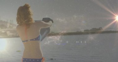 Kadın elinde sörf tahtası tutuyordu. Kozmik ışıltılar ve merceklerle seyahat etmek için suyun kenarında duruyordu. Deniz manzarası, yaz, macera, eğlence, canlı, ufuk, sükunet.