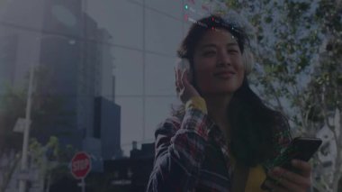 Caddenin köşesinde kulaklıkları ayarlayan, yüzen finans tabloları ve şamdan grafikleri gösteren bir kadın. Kentsel, gündelik, canlı, modern, teknoloji, iletişim, yaşam tarzı