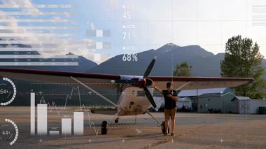 Uçağın yanındaki akıllı telefonu kontrol eden adam, gökyüzünü kaplayan turta ve bar çizelgeleriyle uçuş verilerini gösteriyor. Havacılık, teknoloji, analitik, açık hava, macera, navigasyon, ulaşım