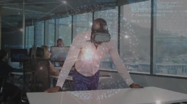 Çalışma masasında VR kulaklık takan bir adam ve çalışma arkadaşları teknoloji ofisinde yüzen kodların arasında yazı yazıyorlar. Gelecekçi, işbirlikçi, yenilikçi, dijital, üretkenlik, gösterişli, profesyonel