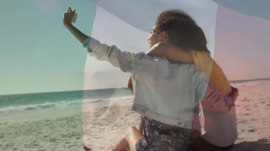 Güneşli sahilde kucaklaşan ve akıllı telefon kullanan çift, yüzen kalp simgeleriyle pazarlamayı gösteriyor. Romantizm, şefkat, eğlence, deniz kenarı, yaşam tarzı, mutluluk, huzur