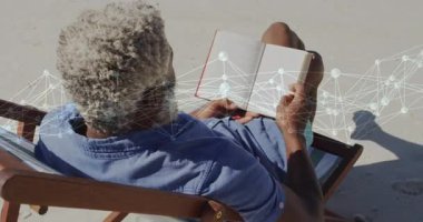 Son sınıf öğrencisi oturmuş, kumsalda ciltli kitap okuyor ve teknoloji ağı düğümlerini gösteriyor. Rahatlama, rahatlama, açık hava, sükunet, sağlık, dijital bütünleşme, modern