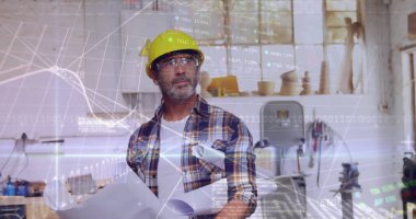 Sarı şapkalı ve gözlüklü bir adam atölyede mali tabloları üst üste dizilmiş planları tutuyor. İnşaat, mühendislik, taslak, endüstriyel, işçilik, tasarım, yenilik