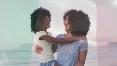 Anne kızını plajda tutuyor, sağlık tablosu ve kalp ikonu gösteriyor. Aile, deniz kenarı, mutluluk, bağ, rahatlık, sükunet, coşku