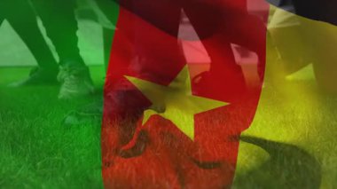 Kamerun bayrağını taşıyan iki oyuncunun bacakları arasında yuvarlanan beyaz futbol topu. Atletizm, sportmenlik, rekabet, açık hava, canlı, takım çalışması, aktif