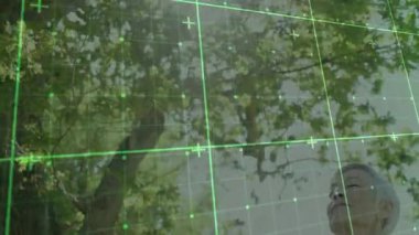 Yapraklı tepe örtüsünün altında, teknolojiyi gösteren yeşil şebeke ile etkileşimde bulunan kıdemli bir kadın. Doğa, büyüme, dinginlik, yenilik, bağlantı, sağlık, farkındalık